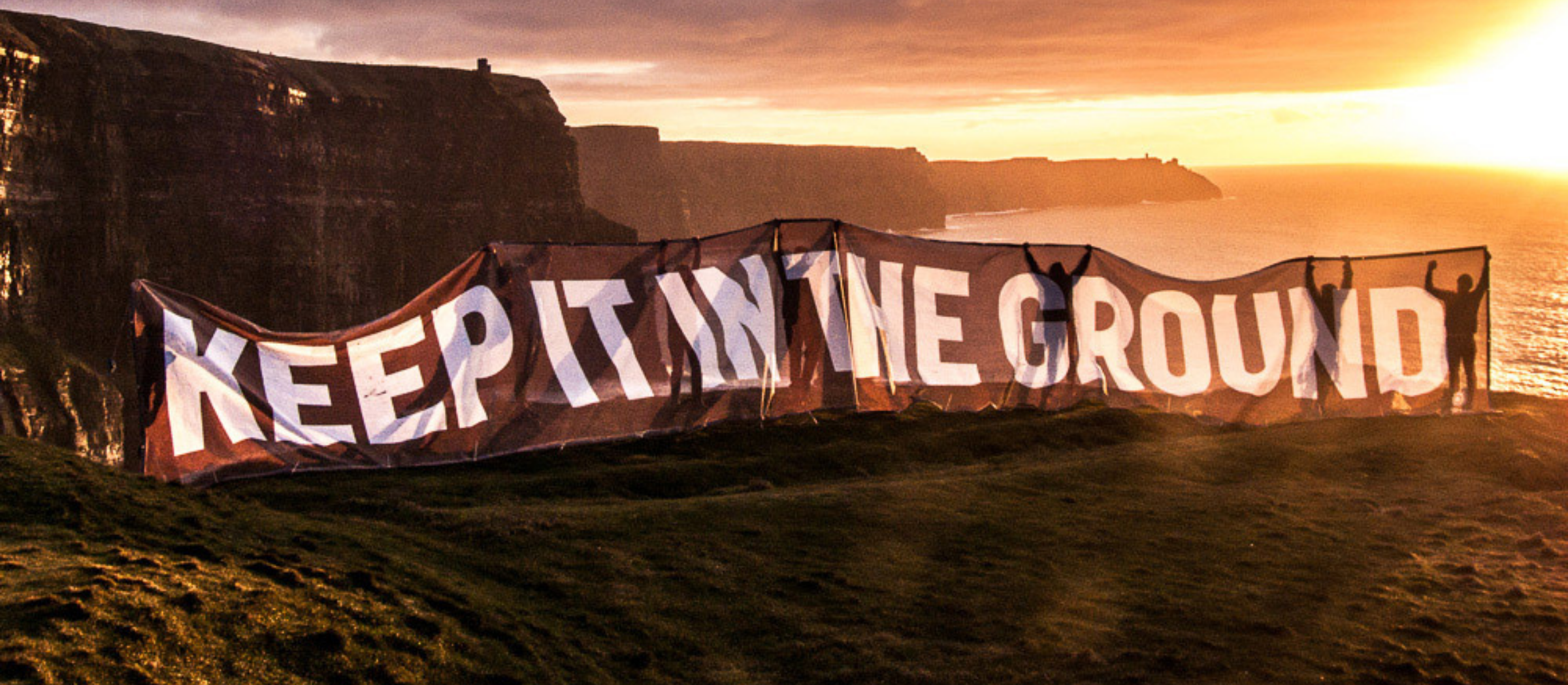 A photo of the sun setting over the Cliffs of Moher. Several people stand in a line holding a large banner that reads, 'Keep it in the ground'