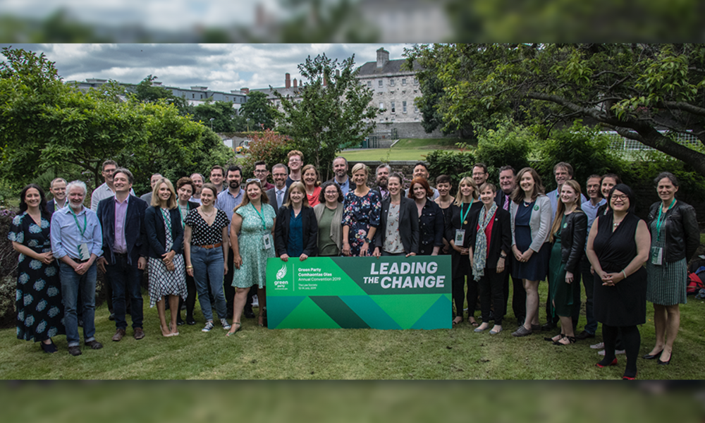 A photo of a large group of people standing outside on a lawn. In front of them is a large sign that reads, 'Green Party Comhaontas Glas Leading the Change'