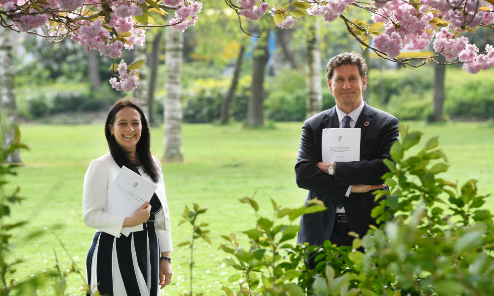 A photo of two people standing in a park, holding A4 paper documents.