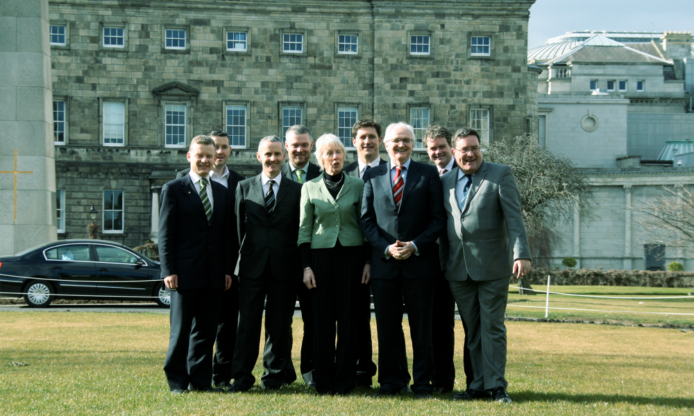 A photo of nine people wearing suits and standing on a lawn in front of a stone building.