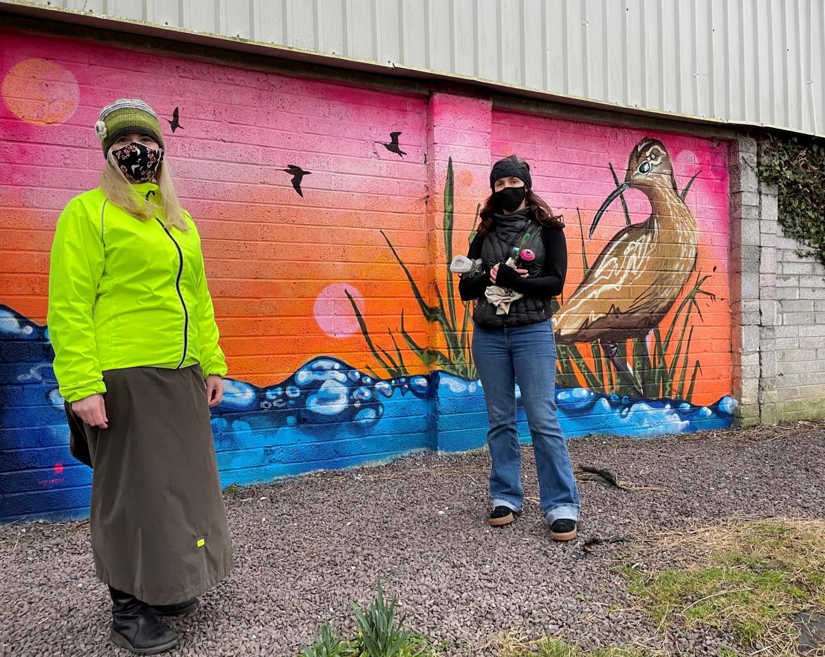 Two people wearing masks stand outside in front of a colourful mural of a curlew. There are oranges, pinks, blues, greens and browns.